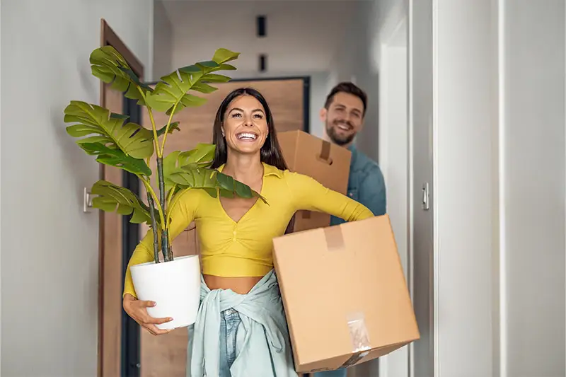 ICS Pack Eine Frau und eine Mann tragen eine Topfpflanze und Umzugskartons in eine Wohnung